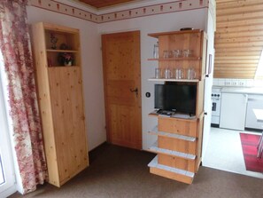 Ein kompakter Wohnraum mit Holztür, Fernseher auf einem Standfuß und Küchenzeile mit weißen Geräten.