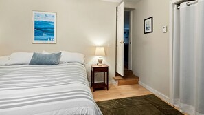 A bedroom with a bed, a nightstand, a lamp, a framed picture on the wall, and a door leading to a hallway. - Lakefront Cabin with Spectacular Sunset Views in Colchester, Vermont (Colchester)