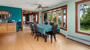 A dining area with a table set for six, wooden chairs, a ceiling fan, and large windows offering a view of greenery.