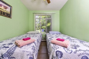 Une chambre avec deux lits, chacun recouvert d’une couvre-lit à motifs floraux et agrémenté de serviettes roses, une fenêtre avec des stores blancs et un ventilateur de plafond.