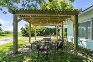 A wooden pergola with a table and chairs underneath, a barbecue grill in the background, and a house with a window to the right. - Pet-Friendly Waterfront Cottage on the Chesapeake Bay, Virginia (Norfolk)