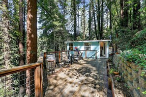A wooden deck leading to a small cabin with a table and chairs, surrounded by tall trees.