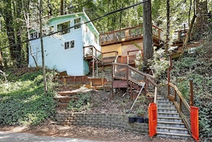 A rustic cabin with a wooden staircase leading to a deck, surrounded by dense forest.