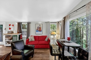 A living room with a red sofa, a leather armchair, a dining table with chairs, a small side table, and a window with curtains.
