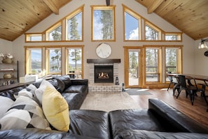 A spacious living room with a fireplace, leather sofas, and large windows offering views of the outdoors. - Exceptional Luxury Cabin with Hot Tub, Games Room, and Fire Pit with Rocky Mountain Views in Grand Lake, Colorado (Grand Lake)