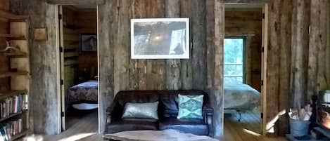A rustic cabin interior with a wooden floor, a leather sofa with cushions, a fireplace, and a bookshelf.