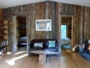 A rustic cabin interior with a wooden floor, a leather sofa with cushions, a fireplace, and a bookshelf.