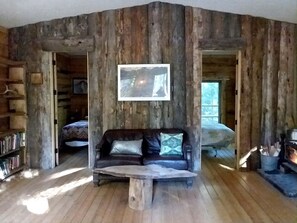 A rustic cabin interior with a wooden floor, a leather sofa with cushions, a fireplace, and a bookshelf.