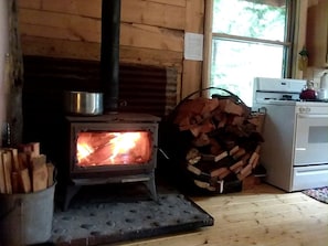 A wood-burning stove, a pot on top, a stack of firewood, and a white oven. - Rustic Woodland Log Cabin with Wood-Burning Stove in Nevada City, California (Nevada City)