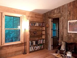 A rustic cabin interior with wooden walls, a bookshelf, a window, and a door leading outside.