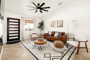 A modern living room with a ceiling fan, a brown sofa, a white armchair, a coffee table, and a large window with curtains. - Dreamy Vacation Rental with Fire Pit and Fantastic Desert Views in Yucca Valley, California (Yucca Valley)