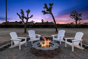 A fire pit with four Adirondack chairs arranged around it, set on a gravel area with desert vegetation in the background. - Dreamy Vacation Rental with Fire Pit and Fantastic Desert Views in Yucca Valley, California (Yucca Valley)