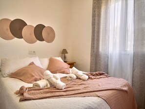 A neatly made bed with pillows, a blanket, and rolled towels. There is a lamp on a bedside table, a wall-mounted light fixture, and a window with sheer curtains. - Las Rosas 6 by Casasol (Frigiliana)