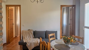 Een gezellige woonkamer met een bank, een eettafel met placemats, een kroonluchter en houten deuren.