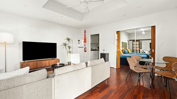 A modern living room with a flat-screen TV, a sofa, a dining area with wicker chairs, and a kitchen in the background. - Cute & Cosy - Walk to Sydney CBD & Harbour (Woolloomooloo)