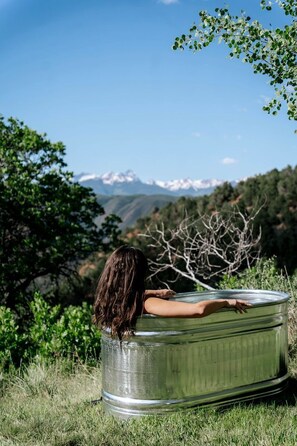 A person sitting inside a large metal tub outdoors with a mountainous landscape in the background.