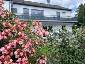 Una casa de dos pisos con un balcón, rodeada de exuberante follaje y flores rosadas en flor.
