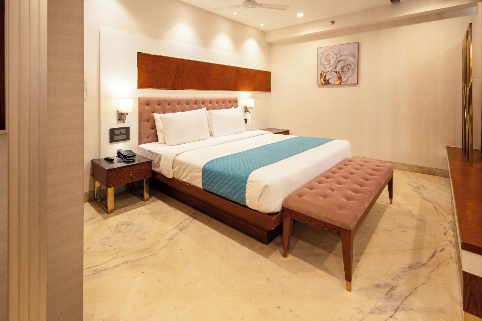 A hotel room with a large bed, a bedside table with a phone, a nightstand, a bench, and a wall-mounted artwork.