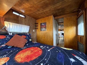 A bedroom with a bed featuring a space-themed bedspread, wooden walls, a window with curtains, and a door leading to another room. - Retro Trailer Rental for Glamping Getaway in Joshua Tree, California (Joshua Tree)