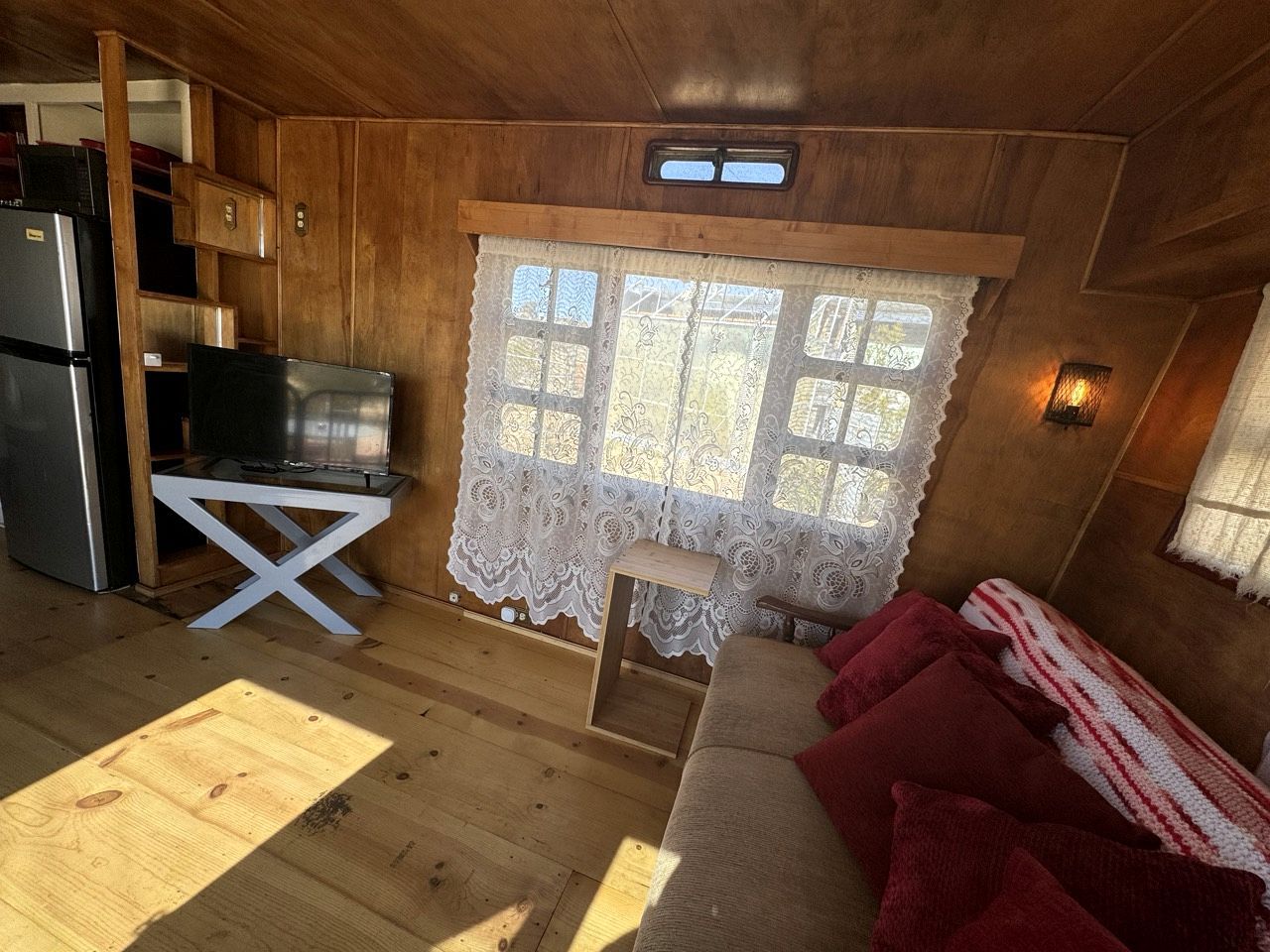 Een interieur van een blokhut met een houten vloer, een kitchenette met een koelkast en fornuis, een eettafel en een raam met kantgordijnen.