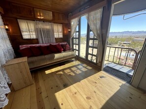 A wooden-floored room with a sofa and curtains, a view of the outdoors, and a door leading outside. - Retro Trailer Rental for Glamping Getaway in Joshua Tree, California (Joshua Tree)