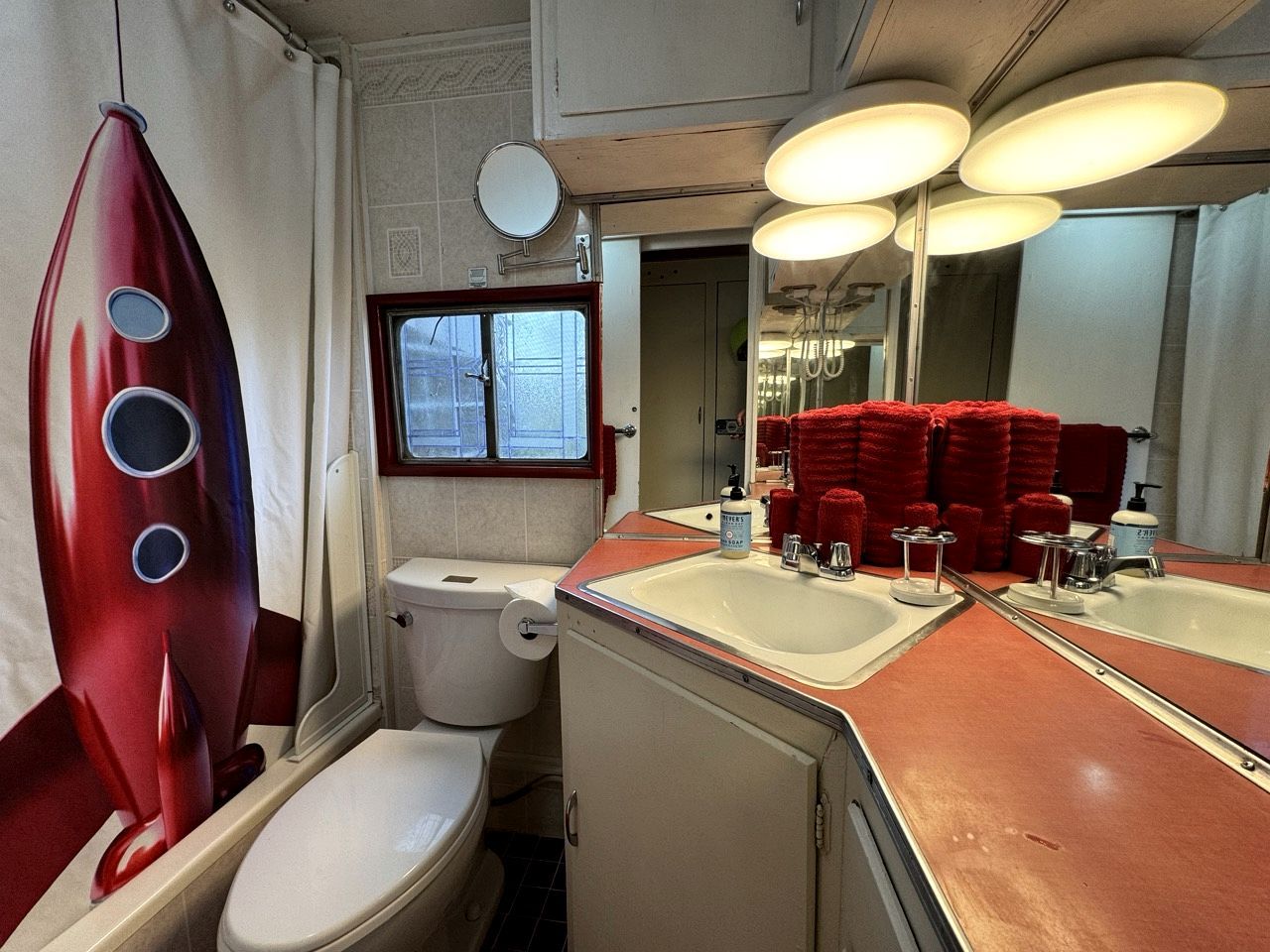 A compact bathroom with a toilet, two sinks, a mirror, and a red towel rack.