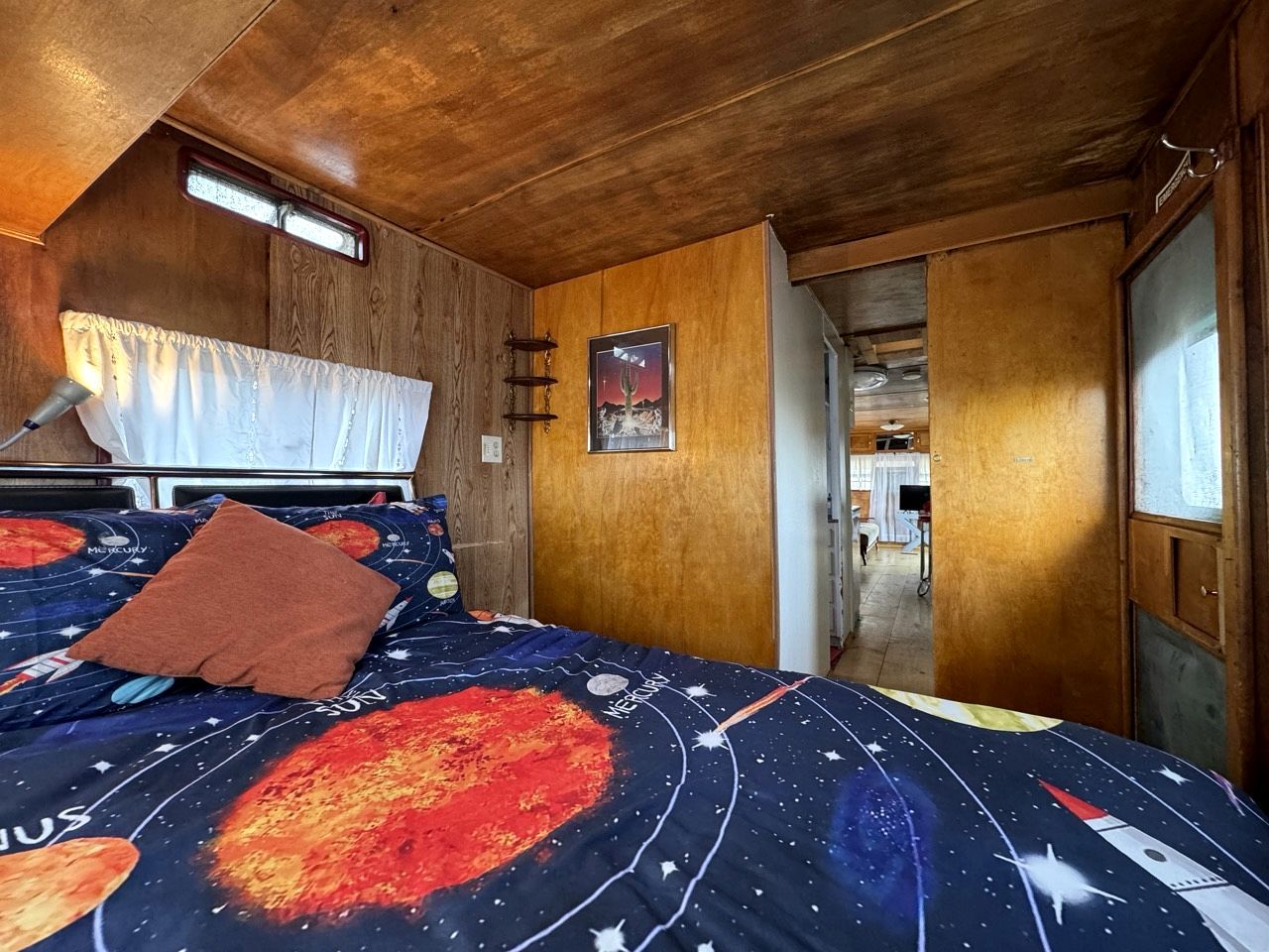 Une chambre à coucher avec un lit recouvert d’une couette à motifs spatiaux, des murs en bois, une fenêtre avec des rideaux et une porte donnant sur une autre pièce.