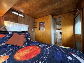 A bedroom with a bed featuring a space-themed bedspread, wooden walls, a window with curtains, and a door leading to another room. - Retro Trailer Rental for Glamping Getaway in Joshua Tree, California (Joshua Tree)
