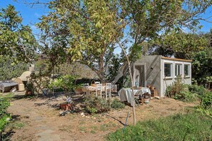 A rustic outdoor setting with a small wooden structure, a table with chairs, and a variety of plants and trees. - Oak Farm Guesthouse Junior (Andros)