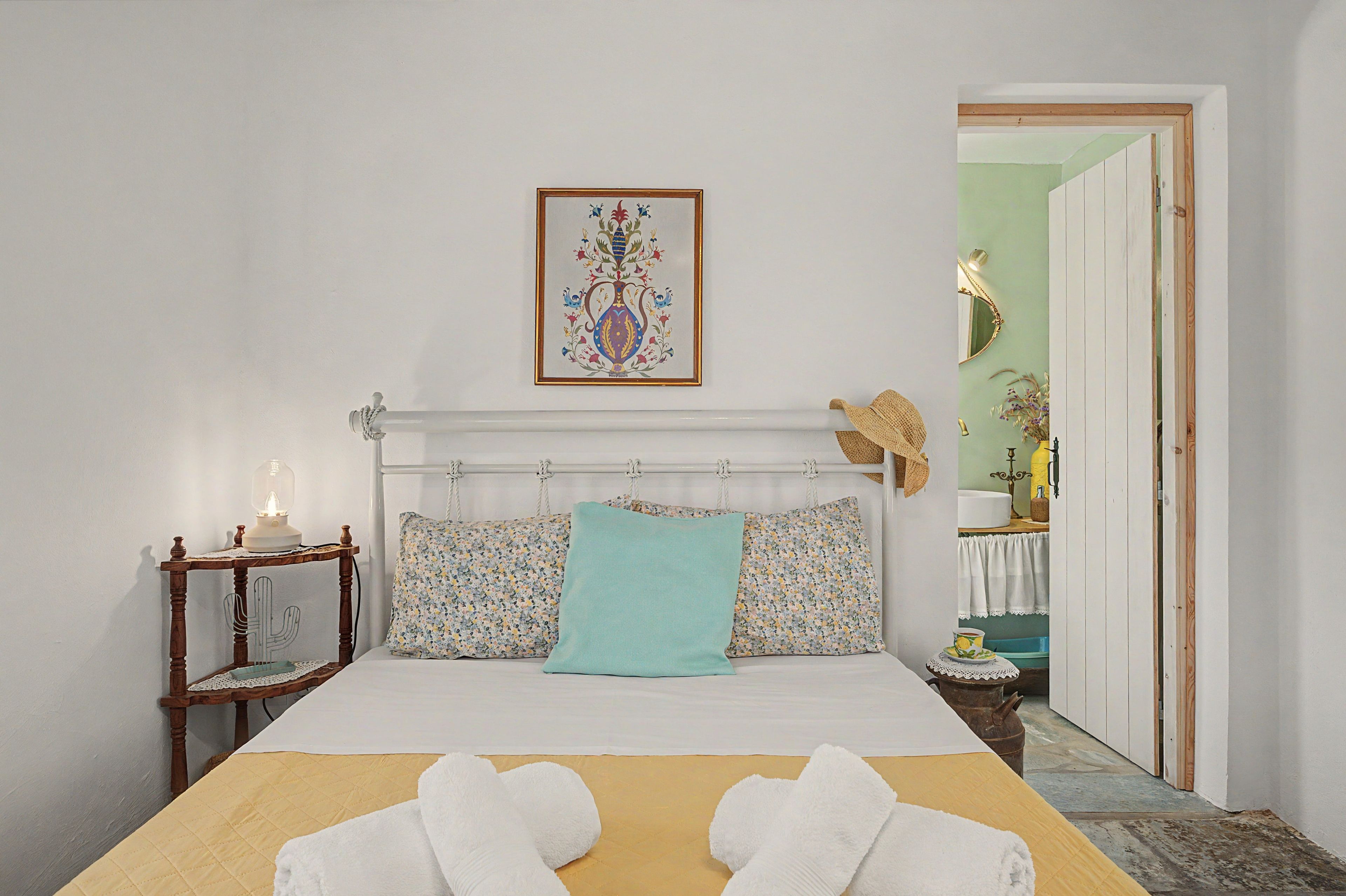 Una cama bien hecha con edredón blanco y dos toallas blancas, una mesita de noche con lámpara, y una obra de arte enmarcada sobre la cama.