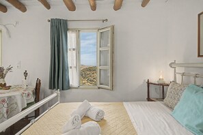 A bedroom with a bed, a window with curtains, a bedside table with a lamp, and a view of the outdoors. - Oak Farm Guesthouse Junior (Andros)