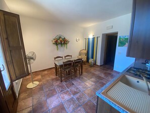 A dining area with a table and chairs, a fan, a decorative wall piece, and a kitchen with a gas stove. - Casa Vacanze Etrusco – Leccio Apartment (San Gimignano)