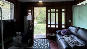 Une pièce de taille modeste, comprenant un canapé, une petite table, un banc, une porte donnant sur l’extérieur et un ventilateur de plafond.