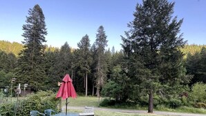 Un espace extérieur verdoyant avec une table et des chaises, un parasol rouge et de grands arbres en arrière-plan.