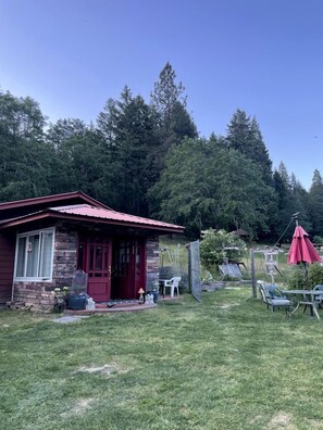 Ein kleines Haus mit roter Tür und Steinaußenwand, umgeben von einem grasbewachsenen Hof und Bäumen.