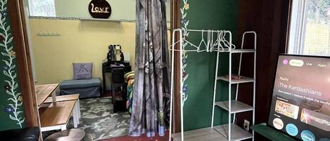A room with a green wall, a patterned rug, a sofa, a TV displaying 'The Kardashians', and a metal rack with clothes.
