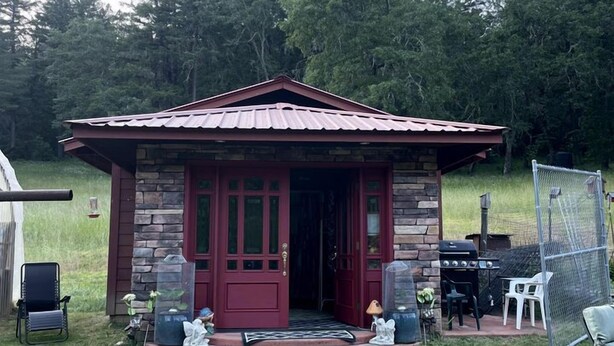 Un petit bâtiment en pierre avec une porte rouge, un porche avec des chaises et un barbecue à l’extérieur.