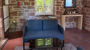 A stone-walled room with a blue sofa, a television on a wooden stand, and a window with a view of greenery.