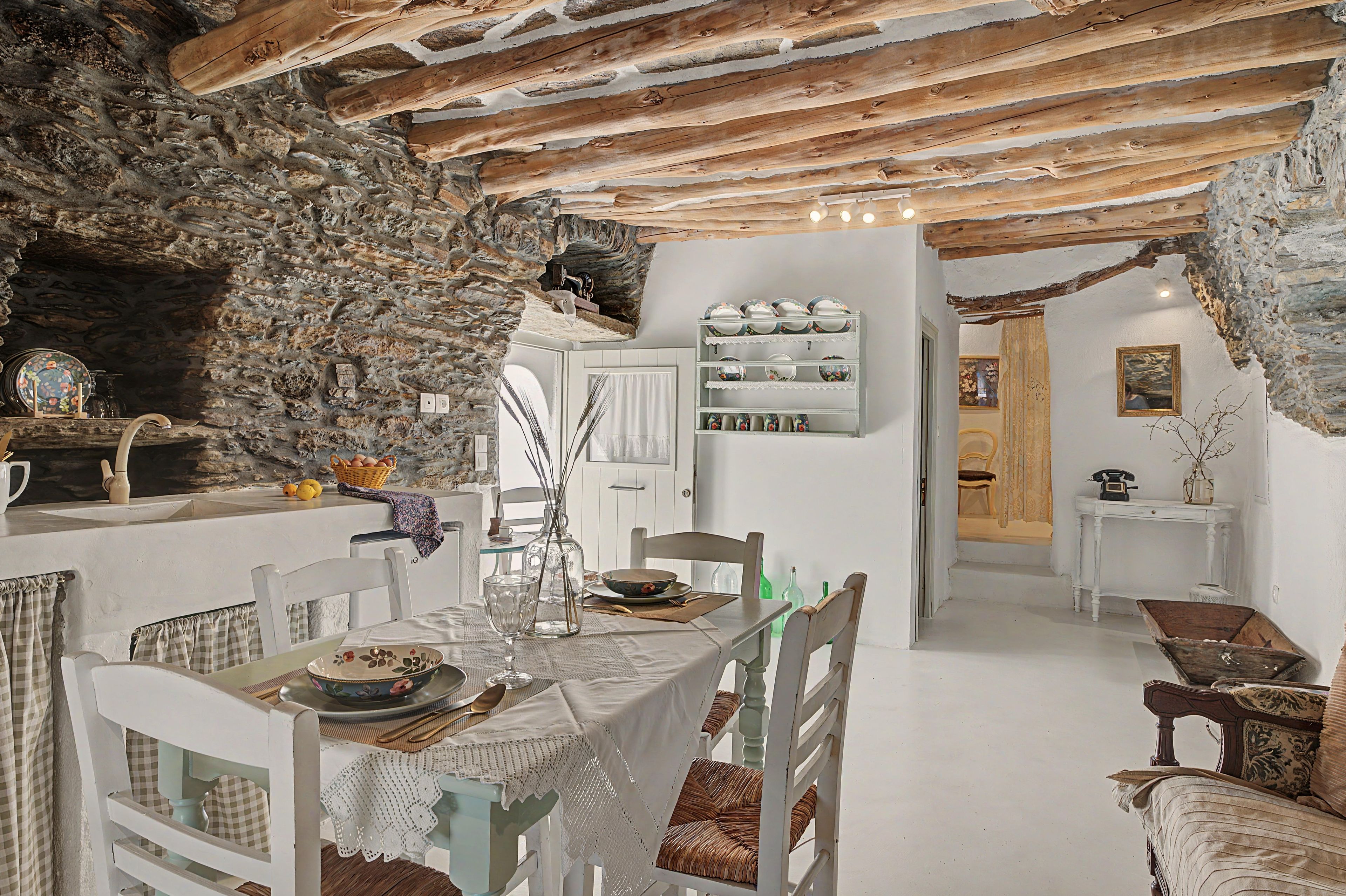 A rustic dining area with a stone wall, wooden beams, and a white table set with plates and glasses.
