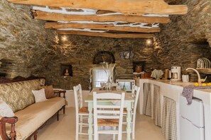 A rustic stone interior with a dining table, white chairs, a sofa, a stone fireplace, and a kitchen area.