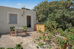 A small, single-story building with a patio area featuring a table and chairs, a wooden fence, and a garden with grapevines.