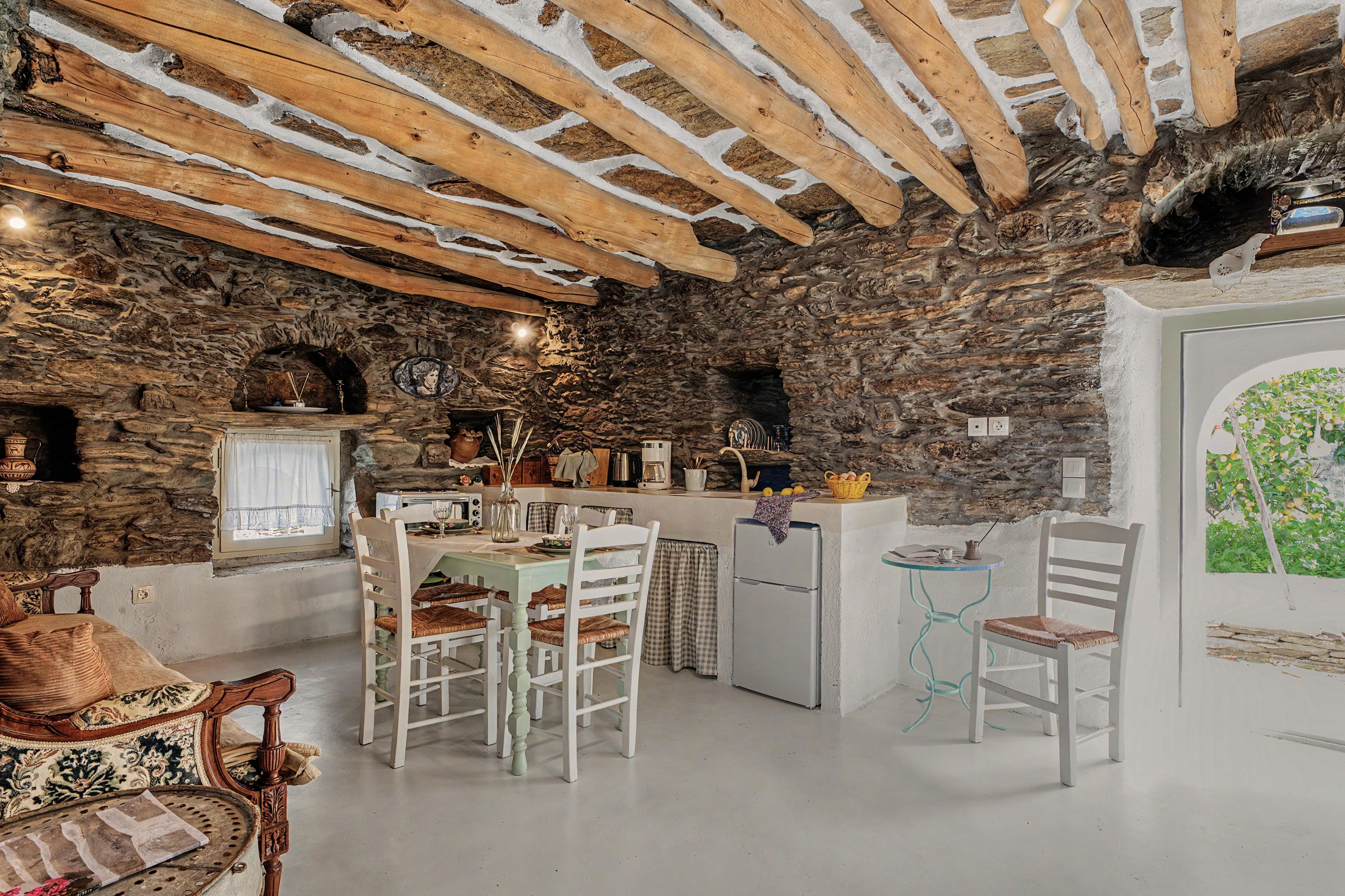 Ein rustikales Interieur mit freiliegenden Steinwänden, Holzbalken, einem Essbereich mit weißen Stühlen und einer kleinen Küche mit Kühlschrank.