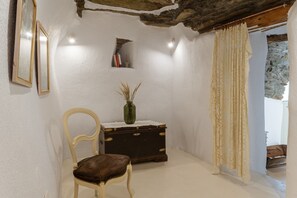 Una habitación con techo rústico, una silla de madera, una mesita con un jarrón, un cofre de madera y una cortina.