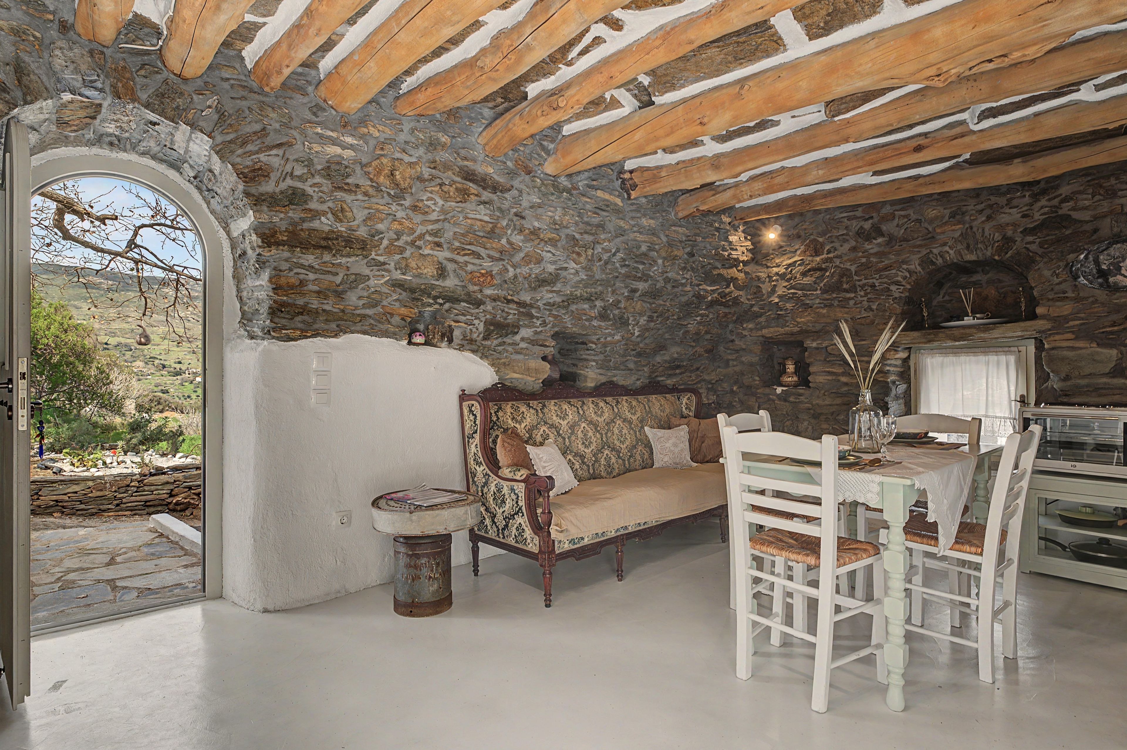 A rustic interior with a stone wall, a wooden ceiling, a sofa, a dining table with chairs, and a view through an archway.