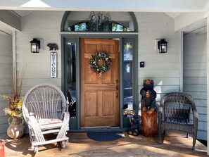 A wooden front door with a welcome sign, two wicker chairs, a potted plant, and a small wooden sculpture.