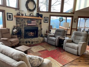 A cozy living room with a stone fireplace, comfortable seating, and a view of mountains through large windows.