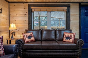 A wooden cabin interior with a leather sofa, two pillows with bear designs, a lamp, and a blue door. - Smoky Mountain Cabin Retreat with Hot Tub, Game Room, Mini Golf & Creek Views in Pigeon Forge, Tennessee (Pigeon Forge)