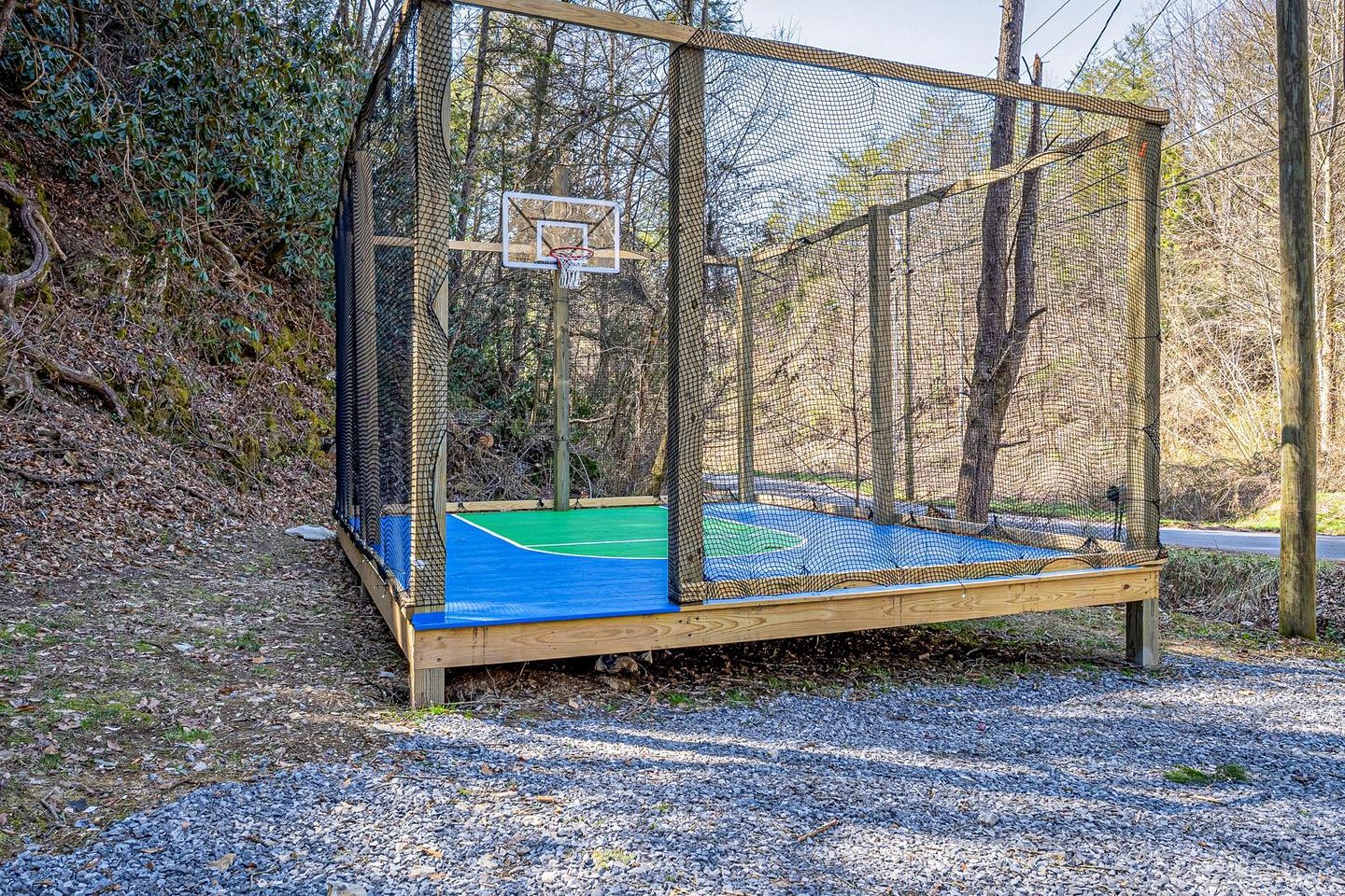 A basketball court with a netted enclosure, surrounded by trees and gravel.