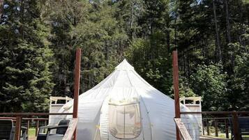 A white tent with a wooden staircase leading up to it, surrounded by a wooden deck and dense forest.