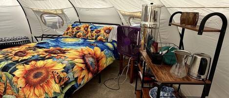 A tented room with a bed, a shelf with kitchen items, and a trash bin.